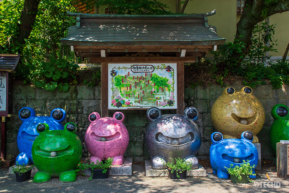如意輪寺入り口に並ぶカラフルなかえる像たち。にっこり笑顔が、訪れる人の心を和ませてくれます。背景には境内案内図があります。