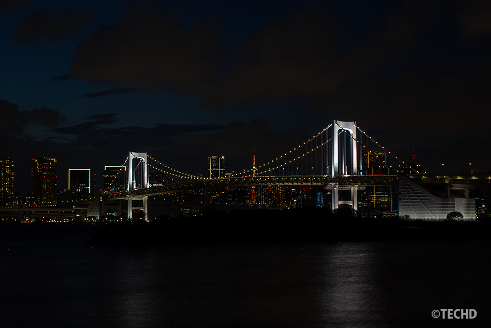 お台場海浜公園展望デッキから撮影したレインボーブリッジの夜景。東京タワーと都心の高層ビル群、ライトアップされた橋が暗い空と水面に映えている。