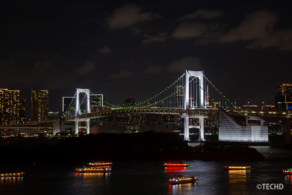 アクアシティお台場 展望テラスから撮影したレインボーブリッジ夜景。緑色にライトアップされた橋と、東京湾を行き交う屋形船の灯りが水面に映えている。