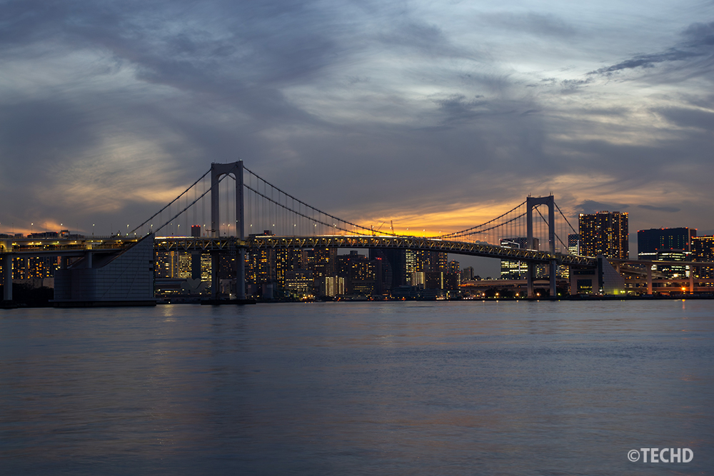 豊洲ぐるり公園から撮影したレインボーブリッジの夕景。雲に覆われた空の下で、夕日の光が少しだけ覗き、橋と都心のビル群を背景に水面に反射している。