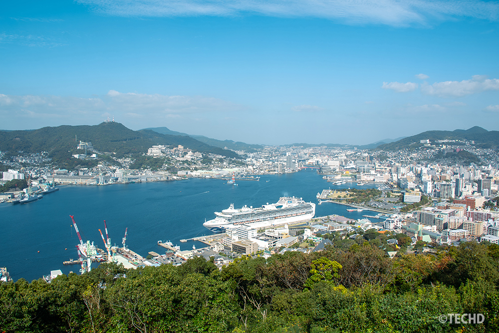 長崎港を見下ろす鍋冠山からの昼景。大型客船と立体的な街並みが湾に沿って広がる様子。