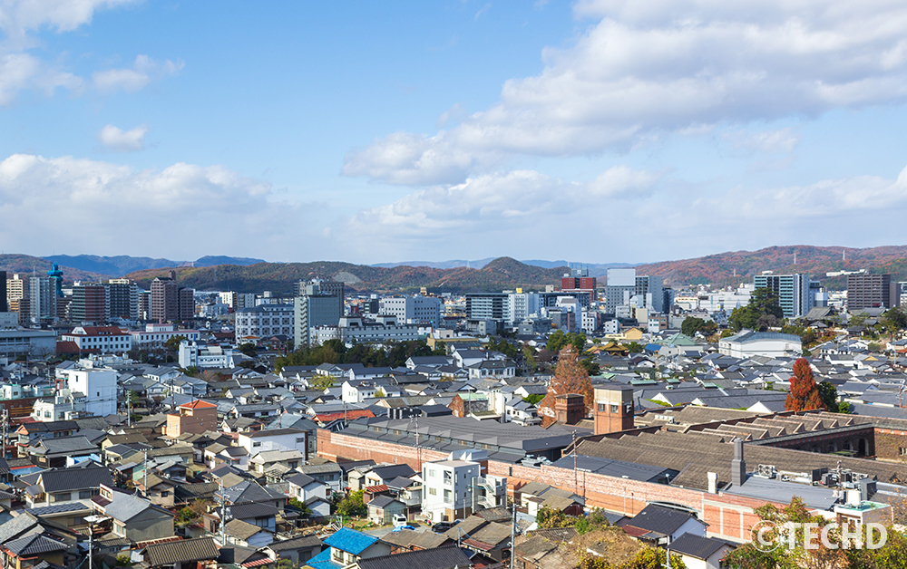 岡山県倉敷市の町並みを小高い丘から一望した風景。伝統的な瓦屋根と現代の建物が混ざり合う、雲の浮かぶ晴天の景観。