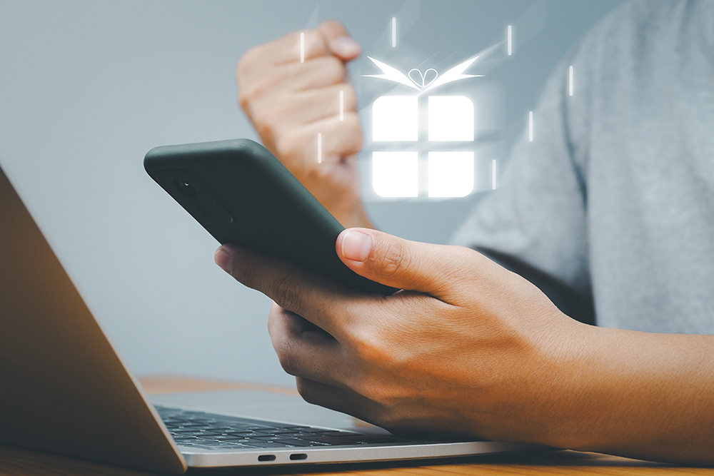 ノートパソコンの横でスマートフォンを手に持ち、もう片方の手でガッツポーズをする人のイメージ。スマートフォン上には光り輝くプレゼントボックスのアイコンが表示されており、クーポン獲得やお得な特典を受け取る喜びを表現している様子。