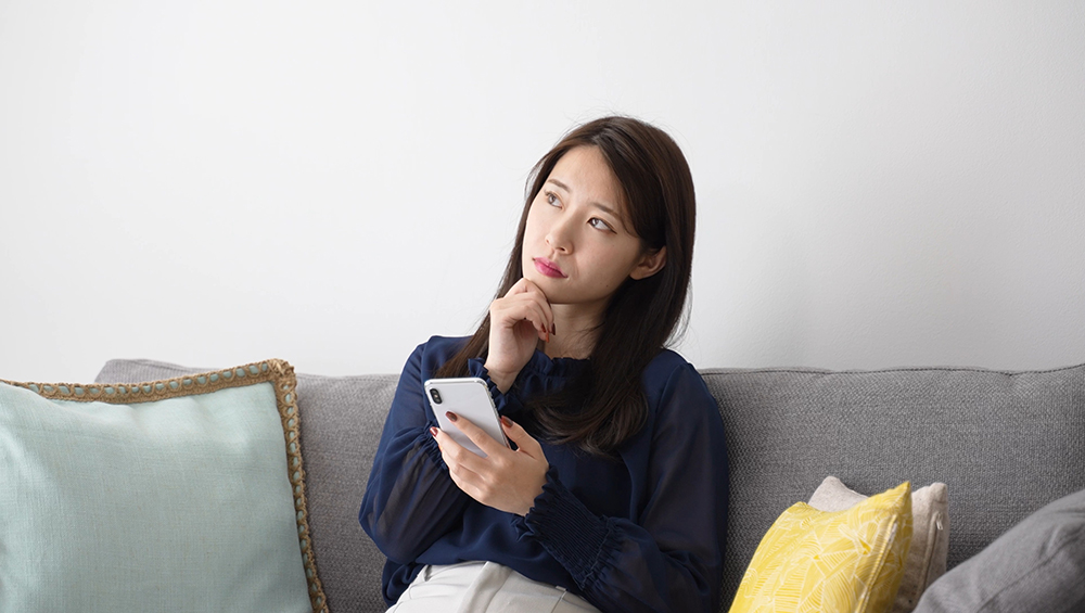 ソファに座り、スマートフォンを手に持ちながら「何を撮ればいいか」と考え込む女性。初心者が抱く撮影テーマへの悩みや迷いを表現したイメージ画像。