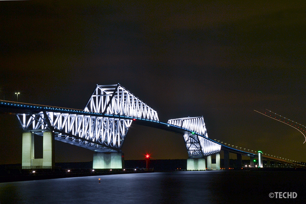 夜の東京ゲートブリッジ。トラス構造が白くライトアップされ、夜空の右側には羽田空港へ向かう飛行機の光跡が二筋の赤いラインとなって美しく描かれている。
