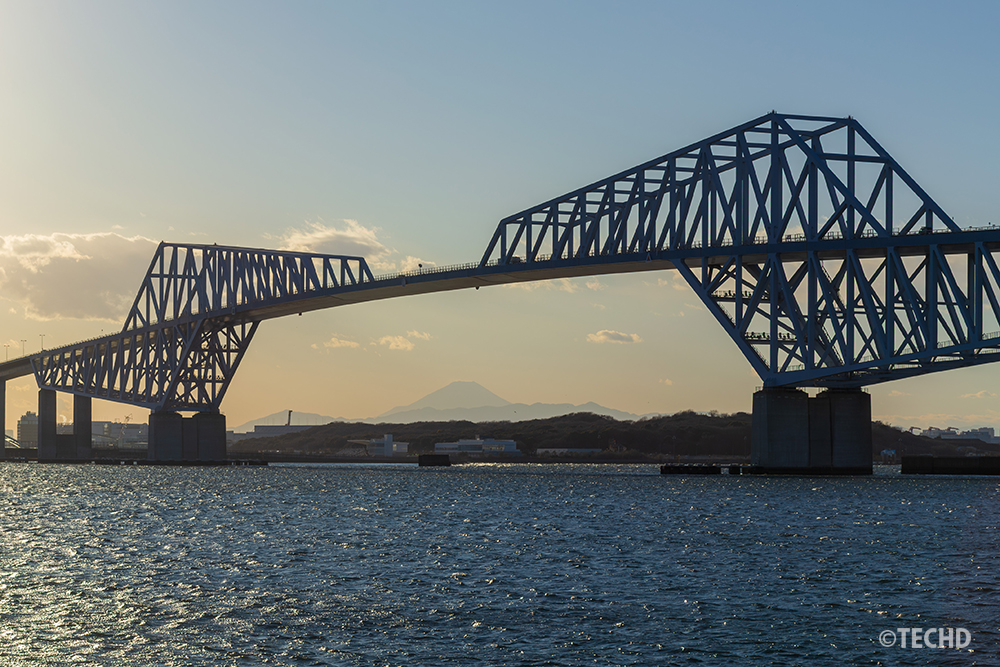 日中の若洲海浜公園から望む東京ゲートブリッジ。トラス構造の橋脚の間に、雪を冠した富士山が重なる現代の絶景構図。