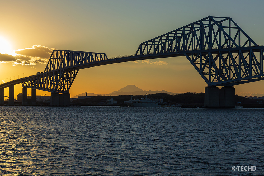 雲の合間から強い夕日が差し込む東京ゲートブリッジと富士山のシルエット。波立つ海面が黄金色に輝く。