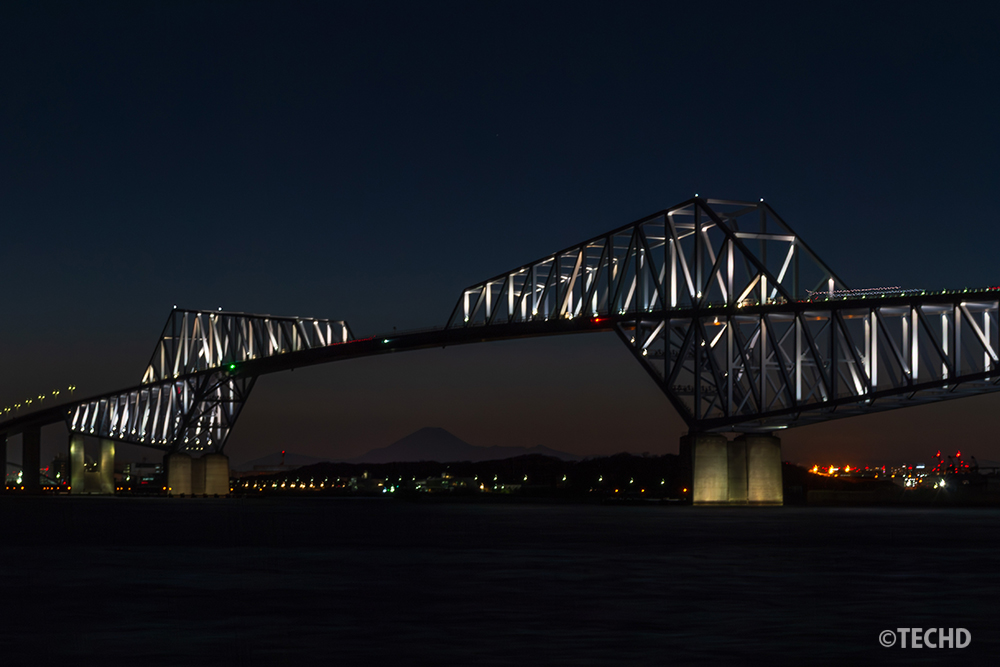 夜の帳が下りた若洲海浜公園から望む、白金色にライトアップされた東京ゲートブリッジ。トラス構造の合間に、薄墨色に溶けゆく幻想的な富士山のシルエットが浮かぶ。