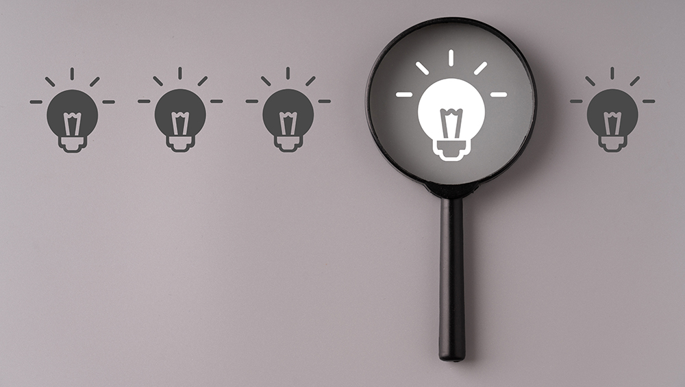 並んだ電球のアイコンの中から、虫眼鏡で照らされた1つの明るい電球が「答え」や「発見」を象徴しているイメージ画像