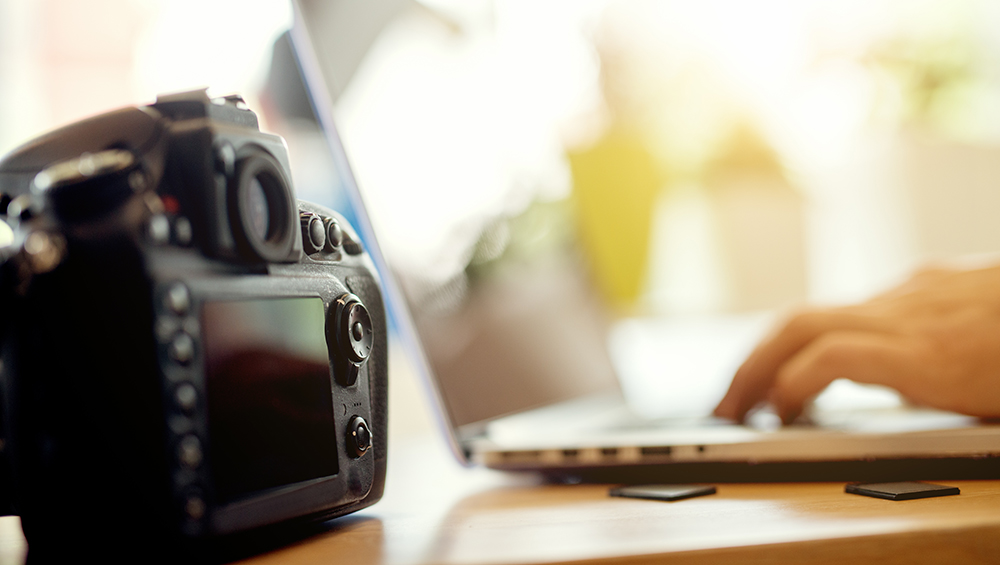 デジタルカメラの背後で、ノートパソコンを使って撮影した写真をストックフォトサイトにアップロードしている作業風景